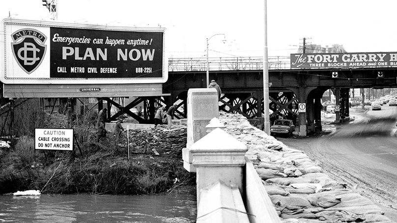 Metro Civil Defence billboard and sandbags at Norwood bridge, 1966. City of Winnipeg Archives, Parks and Recreation Photograph Collection, Box A70, File 2, Item 7.